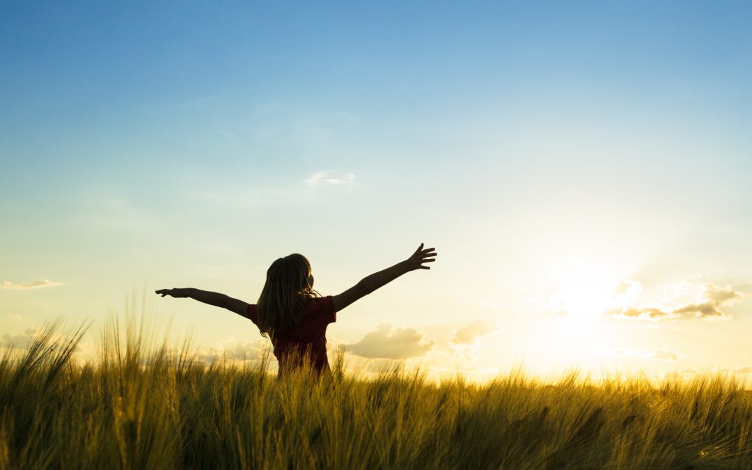 back view of girl stretching her arms out wide, standing in field of tall grasses with sunrising
